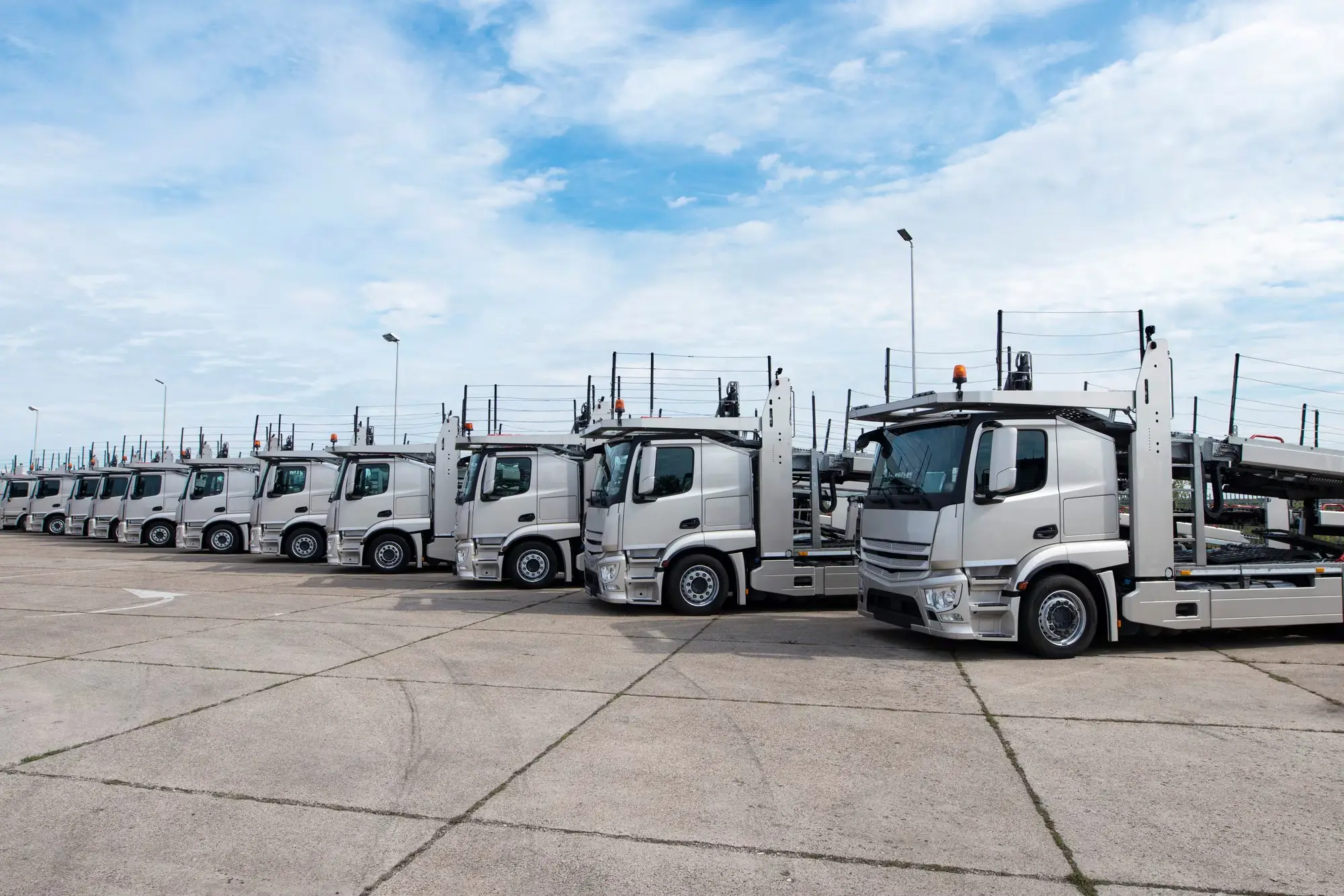 Fleet of silver car carrier trucks parked in a row at logistics yard under blue sky.