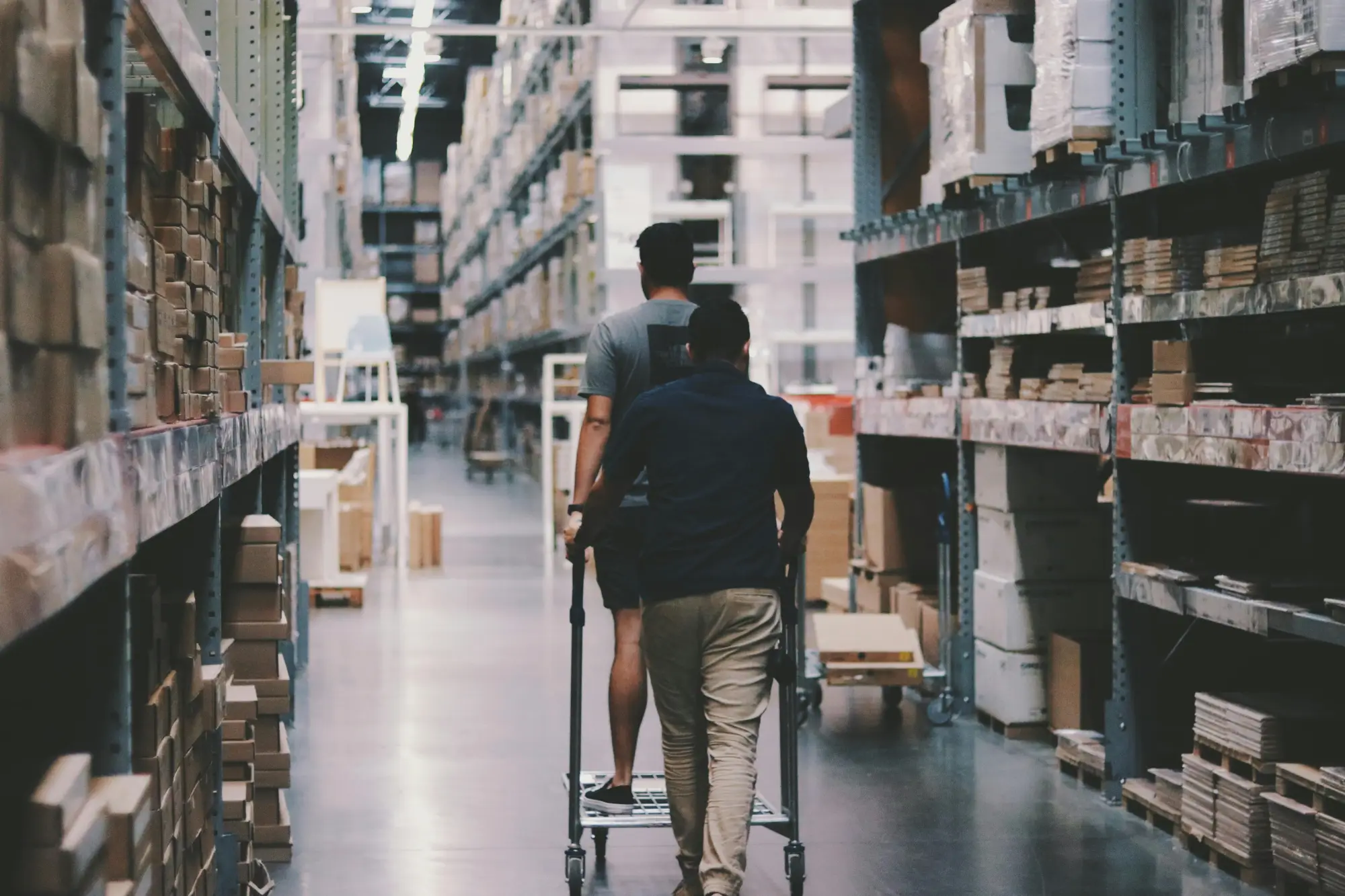 Two people pushing a flatbed cart down a large warehouse aisle lined with shelves of boxed inventory