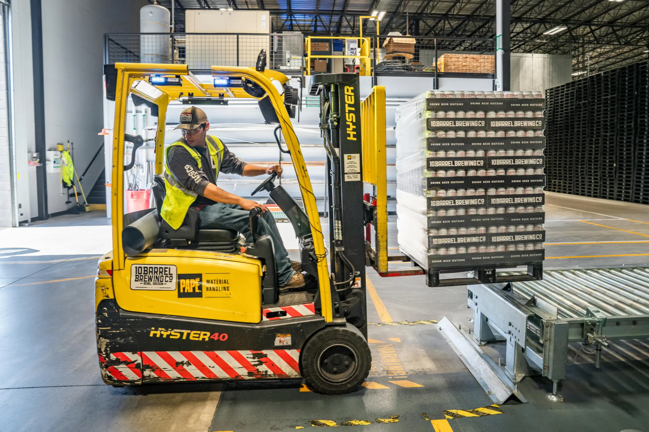 Warehouse employee operating a yellow Hyster forklift to move a pallet of boxed products onto a conveyor line