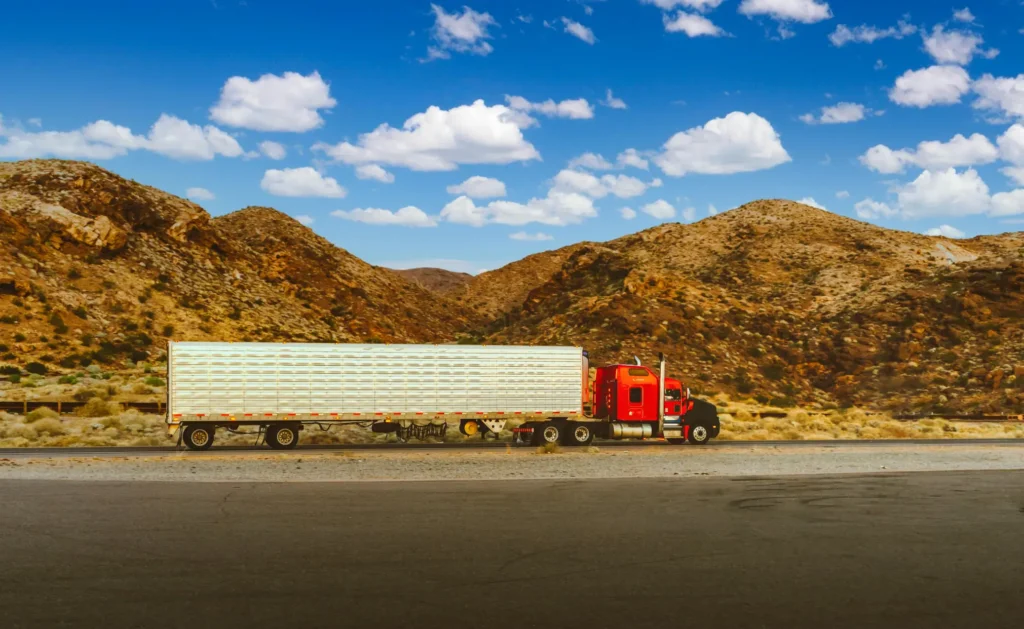 Red semi-truck with a long white trailer driving through a desert highway surrounded by rocky mountains