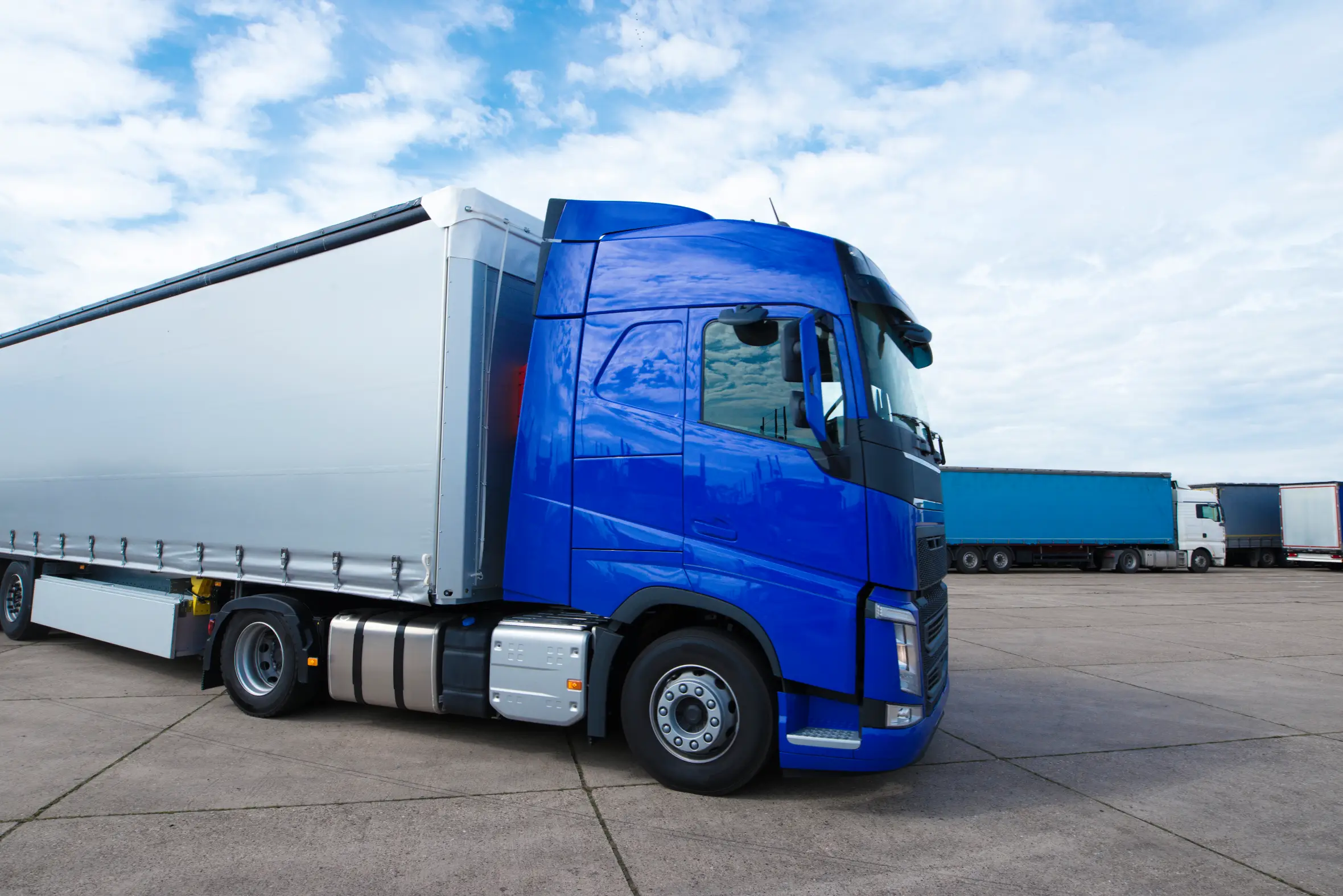 Modern blue semi-truck with a silver trailer parked in an industrial lot under a cloudy blue sky