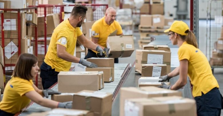 Four warehouse workers in yellow shirts sorting and scanning cardboard boxes on a conveyor belt amid pallet racks and packing stations — 3PL logistics company.